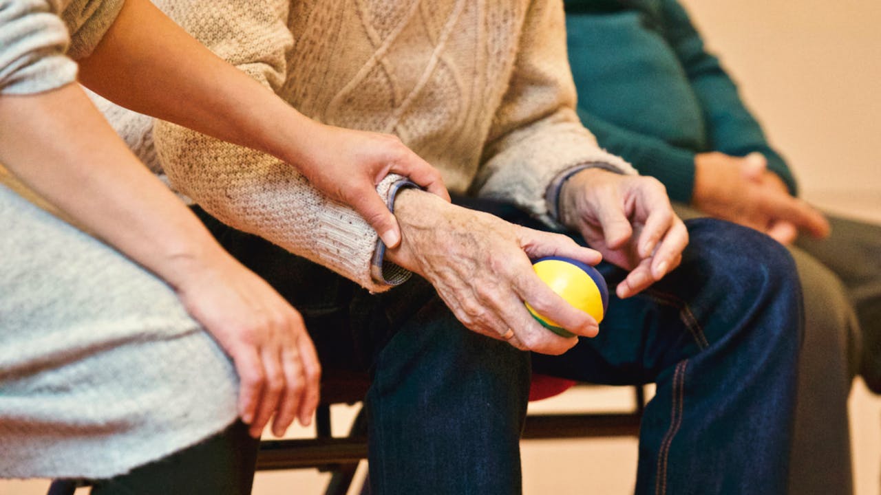 about-01 An elderly person receives support from a caregiver, holding hands indoors, showcasing compassion.
