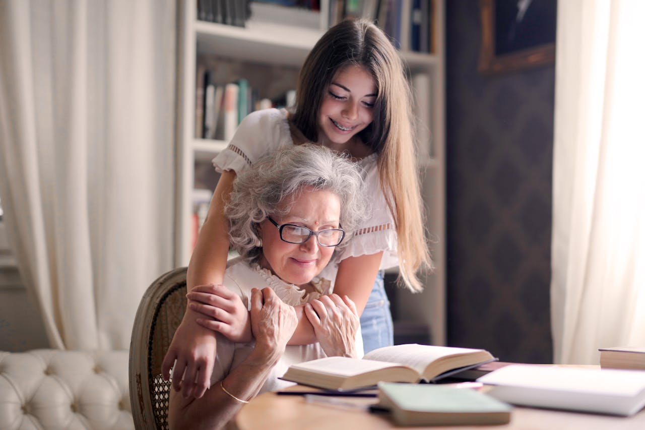 hero-img-02 A heartwarming moment of a granddaughter embracing her grandmother as she reads a book together indoors.