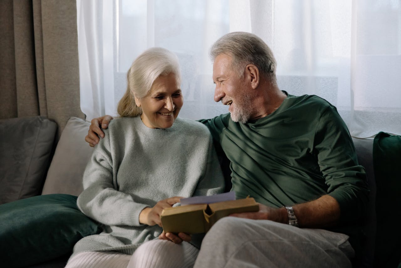 hero-img-01 Senior couple embracing and reading a book on a sofa, sharing a joyful and intimate moment indoors.